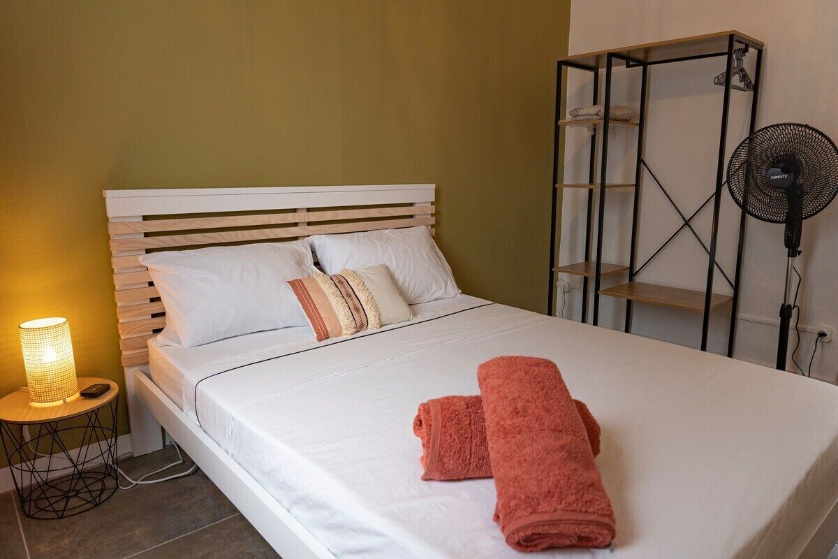 A neatly made bed with white linens and two folded towels, a bedside lamp, and a standing fan.