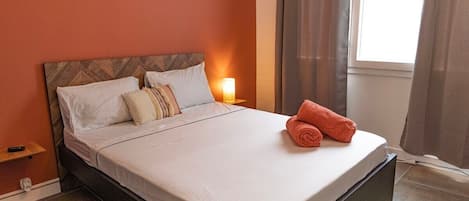 A neatly made bed with white linens and a rolled orange towel, a nightstand with a lamp, and a window with curtains.