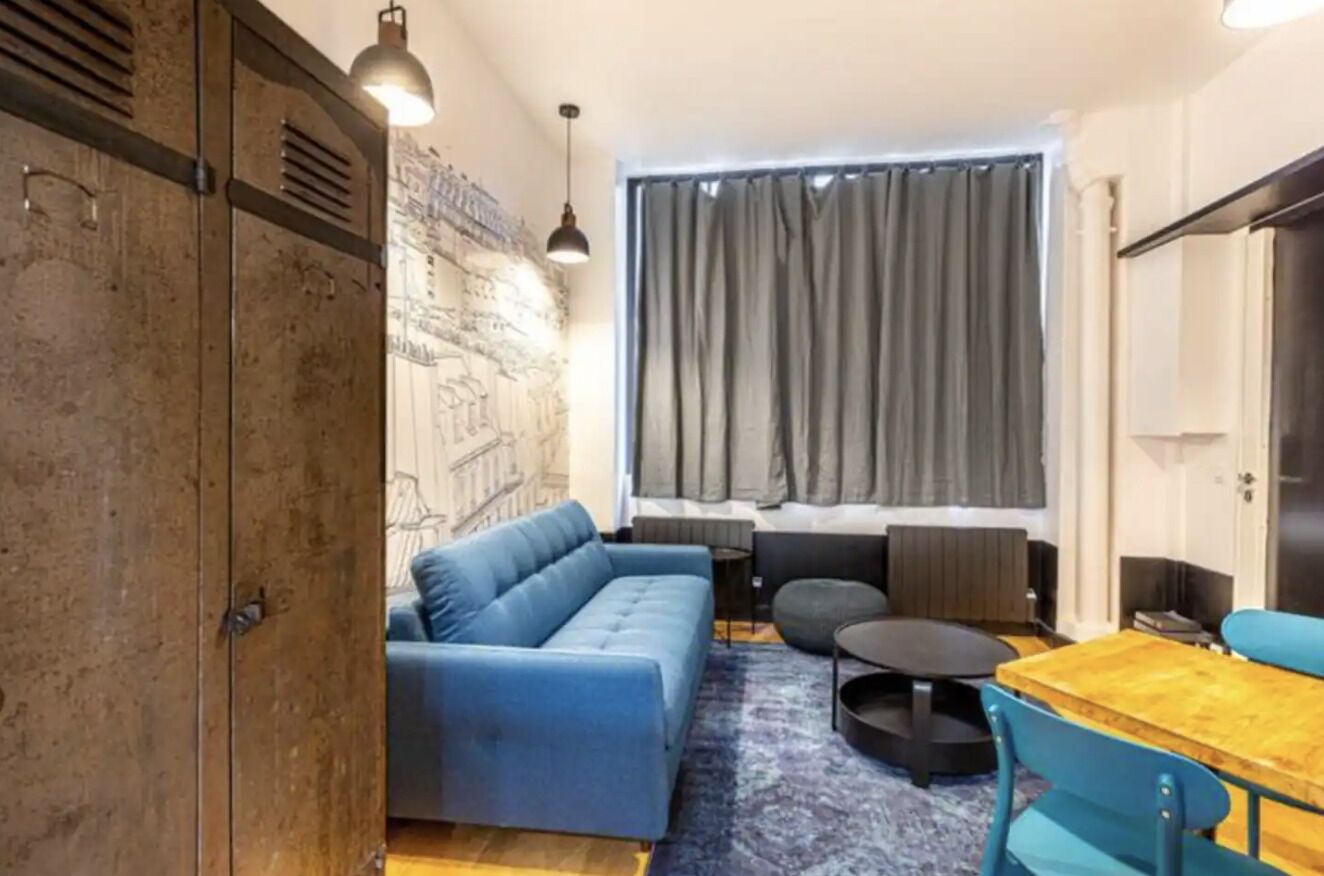 Een moderne woonkamer met een blauwe bank, een houten eettafel en een groot raam met gordijnen.