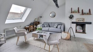 Un salón tipo loft con techo inclinado, tragaluz, sofá gris, una mesa de centro, dos sillones, una lámpara de pie, chimenea y una ventana pequeña.