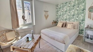 Ein Schlafzimmer mit einem Bett, einem Holztisch, einem Korbstuhl, Wandbehagung aus Makramee und einem Fenster mit Vorhängen.
