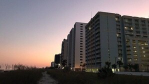 A high-rise hotel with multiple floors and balconies, a pathway leading to the entrance, and a sunset in the background.