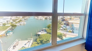 Une vue depuis une fenêtre donnant sur une marina où sont amarrés des bateaux, un quai avec des chaises et des tables, et des bâtiments en arrière-plan.