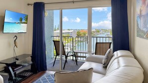 Un salon avec un canapé blanc, un téléviseur fixé au mur, un jeu d’échecs et une vue sur une marina, visible à travers de grandes portes vitrées.