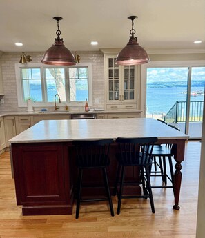 A kitchen with a large island, two pendant lights, a view of the sea, and a dining area with chairs.
