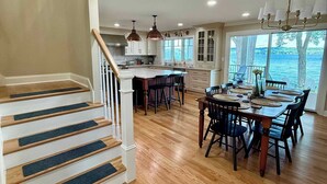 A spacious kitchen with wooden floors, a dining area with a large table, and a view of a lake through large windows. - NEW-Skaneateles Level Lakefront-Ready to Rent! (Skaneateles)