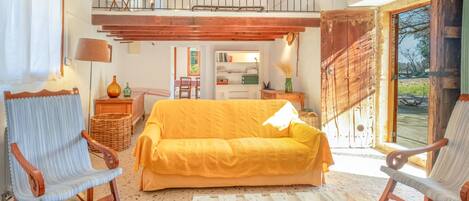 Un amplio salón con un sofá amarillo, dos mecedoras, una mesa de centro con libros y naranjas, y vistas al exterior a través de una gran puerta de madera.