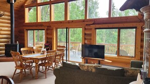 Interior de una cabaña de troncos con comedor, chimenea, televisor y ventanas amplias que ofrecen vistas al bosque.