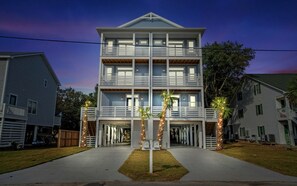 A multi-story building with a balcony on the second floor, a covered entrance, and palm trees in front. - Modern Beach Townhome w/ Hot Tub, Fire Pit, EV Golf Cart & Rooftop Deck (Carolina Beach)