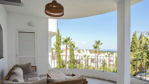 Ein Balkon mit einer Couch, einer hängenden Leuchte und Blick auf das Meer und viel Grün.
