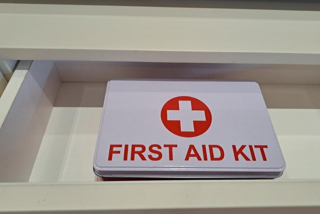 A white first aid kit with a red cross symbol and the text "FIRST AID KIT" on a white shelf.