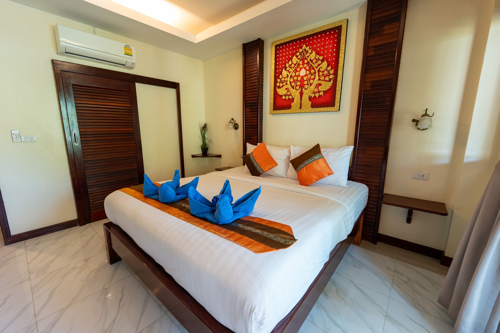 A hotel room with a large bed, a wooden headboard, a wall-mounted air conditioner, a decorative wall hanging, and a small shelf with a plant.