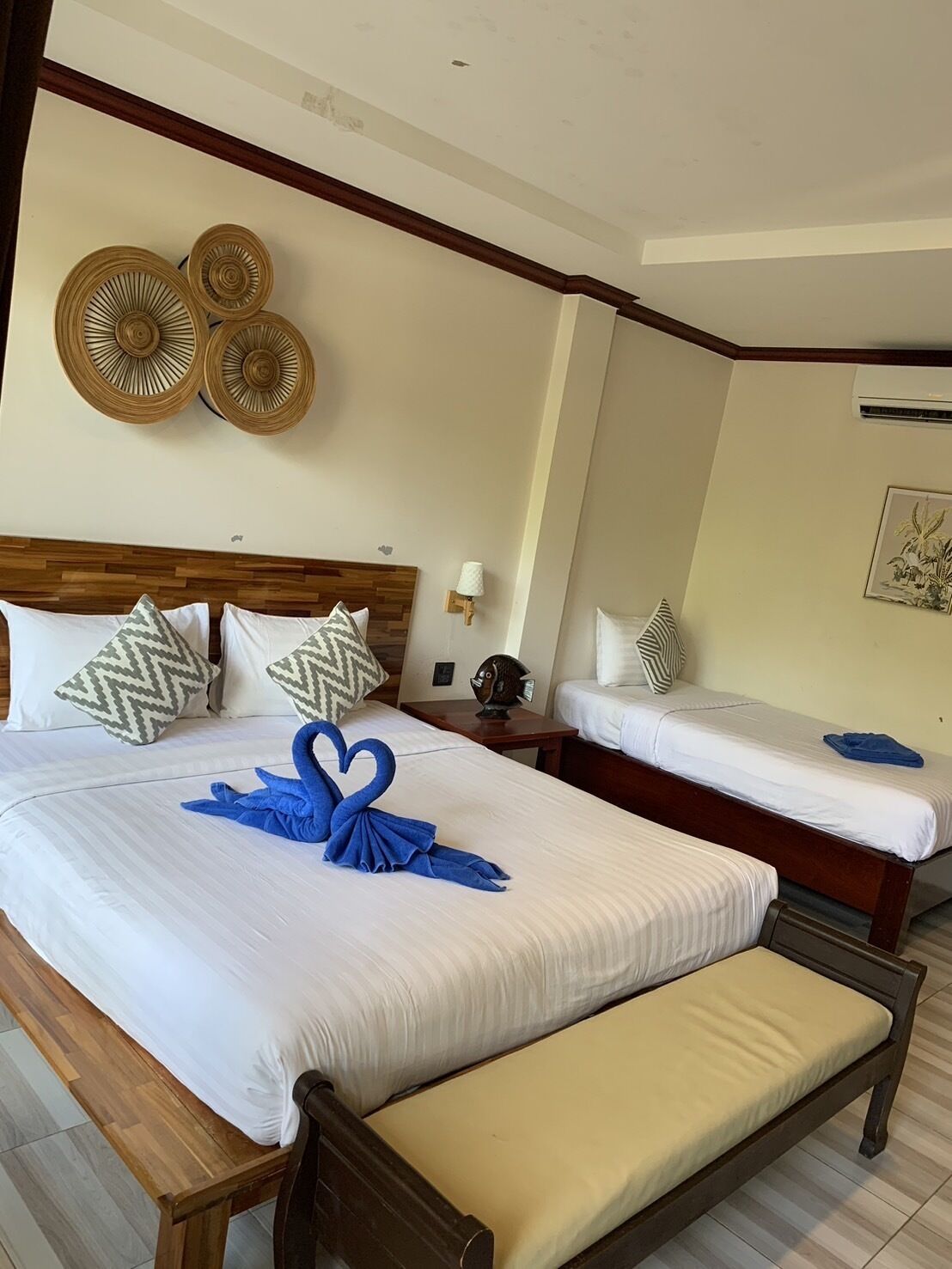 Une chambre d’hôtel avec un grand lit, un lit plus petit, un banc et un climatiseur fixé au mur.