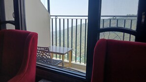 A balcony with two red chairs, a small table, and a view of a hilly landscape. - Hotel Grand Hill (Dharamshala)