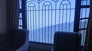 A room with two armchairs and a small table, a large window with a metal railing, and a view of a mountainous landscape. - Hotel Grand Hill (Dharamshala)