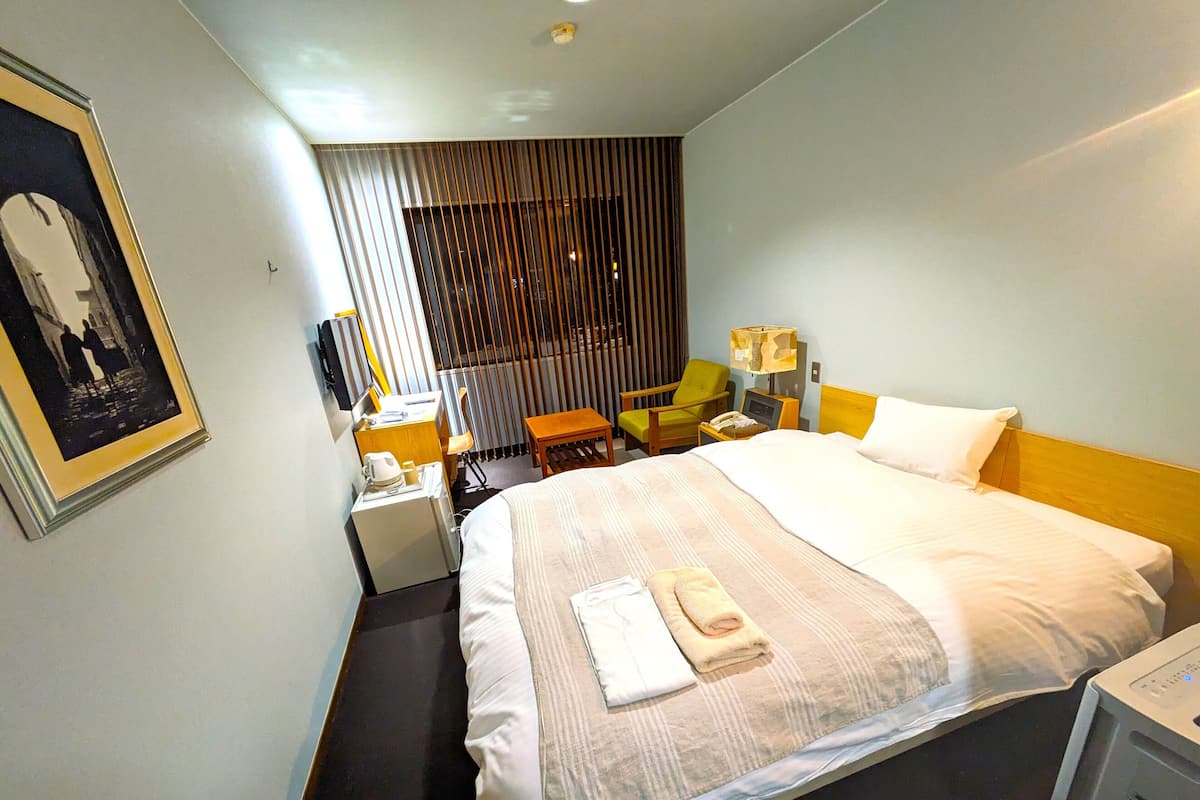 A hotel room with a large bed, a desk with a chair, a small table, a lamp, and a picture on the wall.