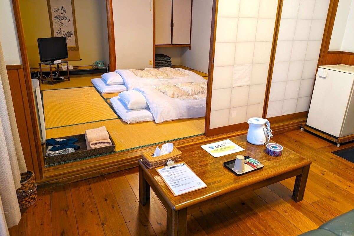 A traditional Japanese-style room with tatami flooring, a bed with white bedding, a wooden table with a teapot, and a small refrigerator.