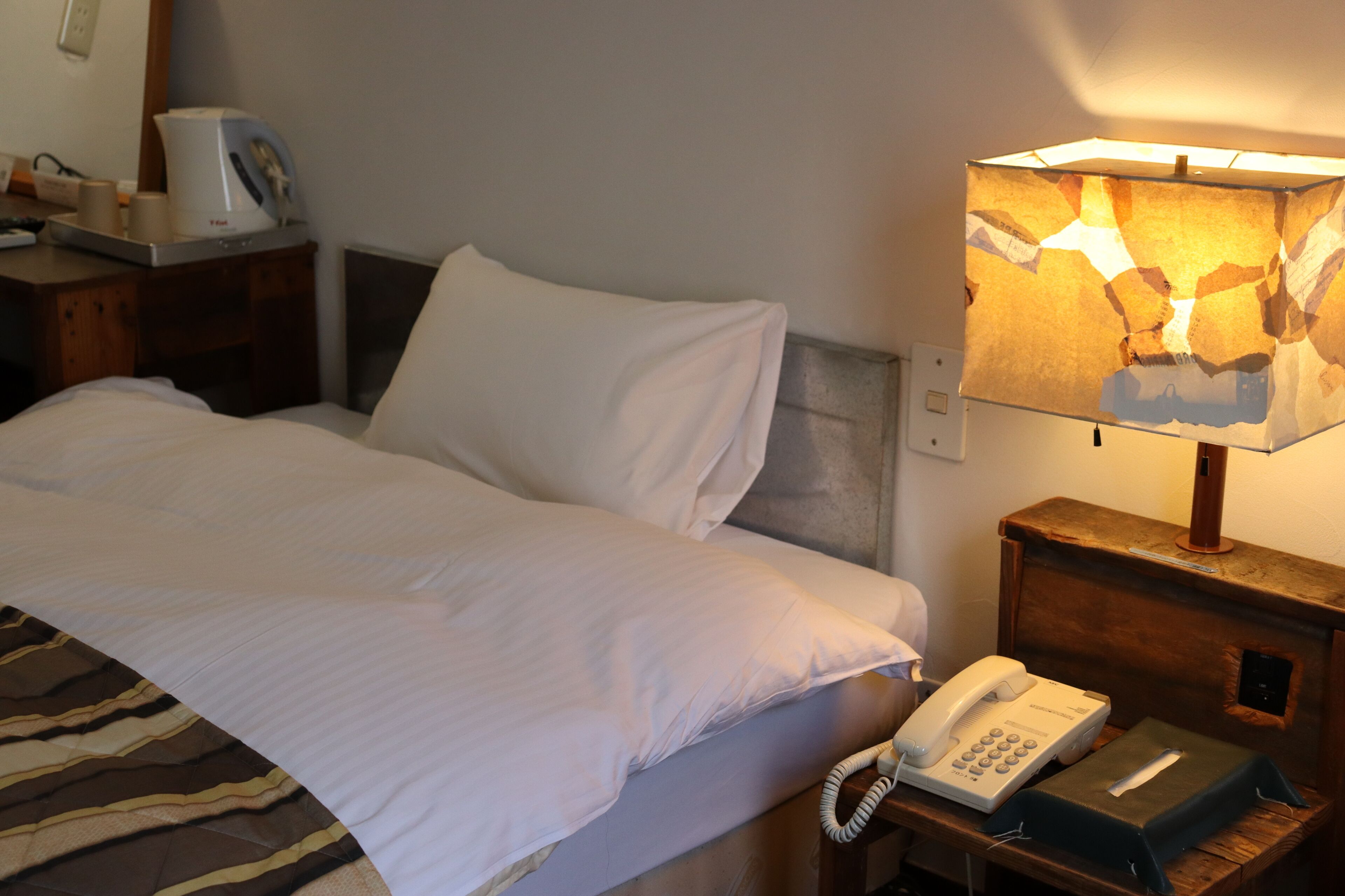 Una cama bien hecha con sábanas blancas, una lámpara de noche, un teléfono y una cafetera sobre una mesita de madera.