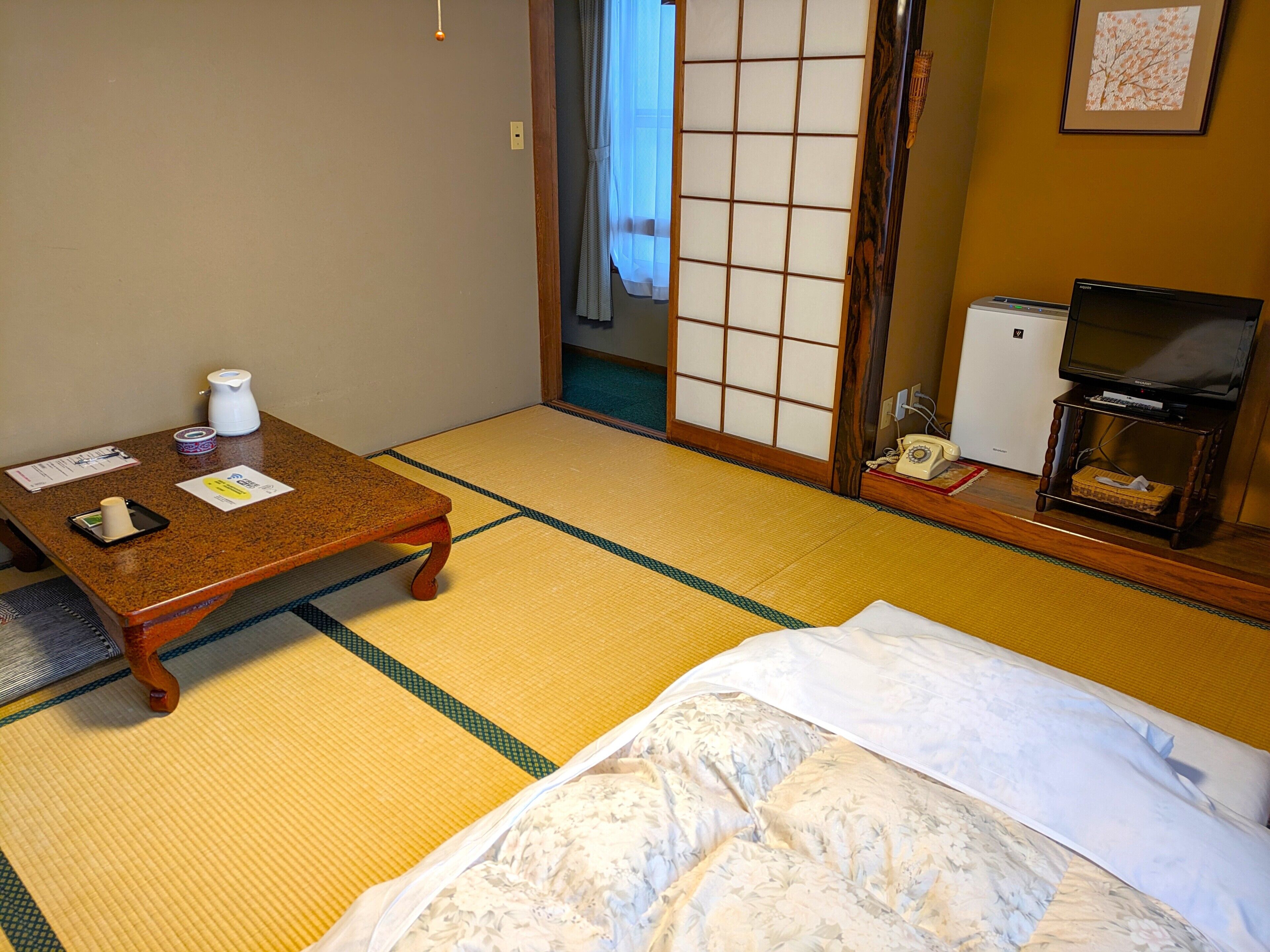 Una habitación japonesa tradicional con suelo de tatami, una mesa de madera baja, una cama con colcha blanca, un televisor sobre un mueble de madera y un refrigerador pequeño.