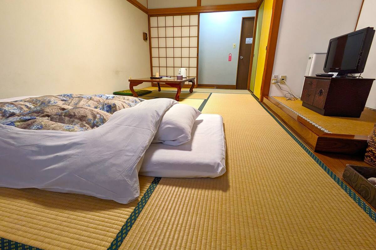 A traditional Japanese room with tatami flooring, a bed with a patterned blanket, a wooden table with a cushion, a television on a cabinet, and a sliding door leading to another room.