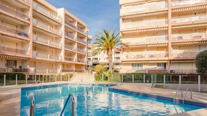 A swimming pool surrounded by multi-story buildings with balconies, a palm tree, and a clear sky.