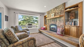 A living room with a stone fireplace, a sofa, a bookshelf, and a view through a large window.