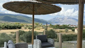 Une terrasse dotée de meubles en osier et d’un grand parasol, offrant une vue imprenable sur un paysage magnifique, avec des montagnes et une étendue d’eau.