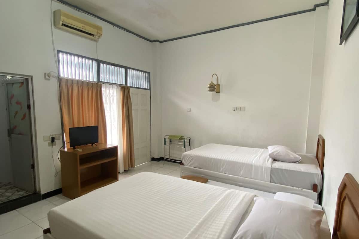 A hotel room with two beds, a TV on a wooden stand, an air conditioner, a window with blinds, and a small table with a lamp.