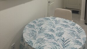 A room with a round table covered with a leaf-patterned tablecloth, a mounted flat-screen TV on the wall, and a white door with a vent. - New amazing location  (Antioch)