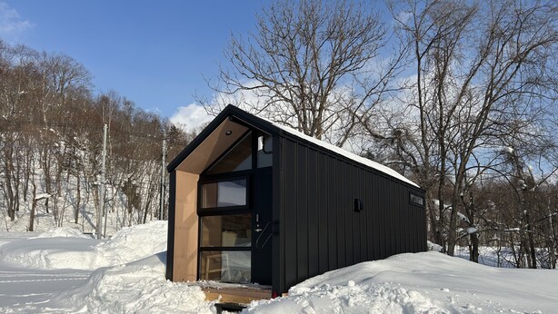 A modern, dark-colored cabin with a large glass door and a wooden interior, surrounded by snow and trees.
