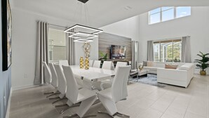 A modern living room with a dining area, featuring a white dining table, chairs, a sofa, and a TV mounted on the wall. - Precious Villa 5Beds/4Baths/Pool/18Min Disney (Davenport)