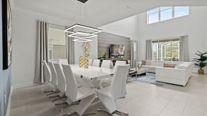 A modern living room with a dining area, featuring a white dining table, chairs, a sofa, and a TV mounted on the wall.