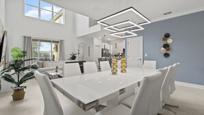 A spacious dining area with a white table and chairs, a modern ceiling light fixture, and a view of the kitchen in the background. - Precious Villa 5Beds/4Baths/Pool/18Min Disney (Davenport)