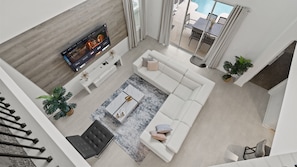 A modern living room with a white sofa, a coffee table, and a flat-screen TV mounted on a wooden wall. There is a staircase leading to an upper level, and a view of a swimming pool through large windows.