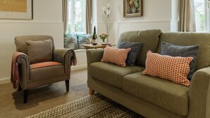 Ein Wohnzimmer mit einer Couch, einem Sessel und einem kleinen Tisch mit einer Flasche und Blumen.
