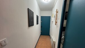 A narrow hallway with white walls, a blue door, and a radiator. There are two framed items on the wall.