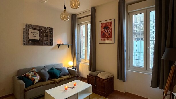 A cozy living room with a sofa, a coffee table, and a lamp. There are windows with curtains and a poster on the wall.