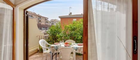 An open balcony with a table set for two, surrounded by white chairs, overlooking a residential area with buildings and greenery.