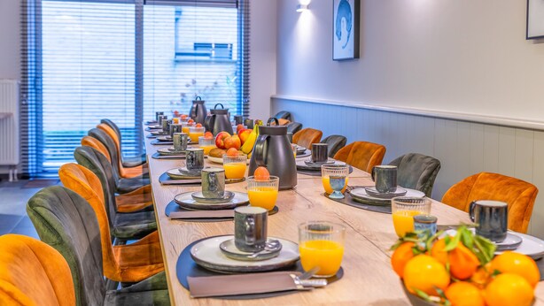 A breakfast setting with a long table, orange chairs, and a bowl of oranges.