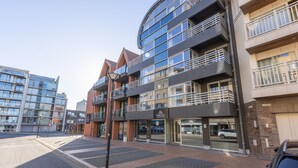 A modern multi-story building with a glass facade, balconies, and a ground-level entrance.