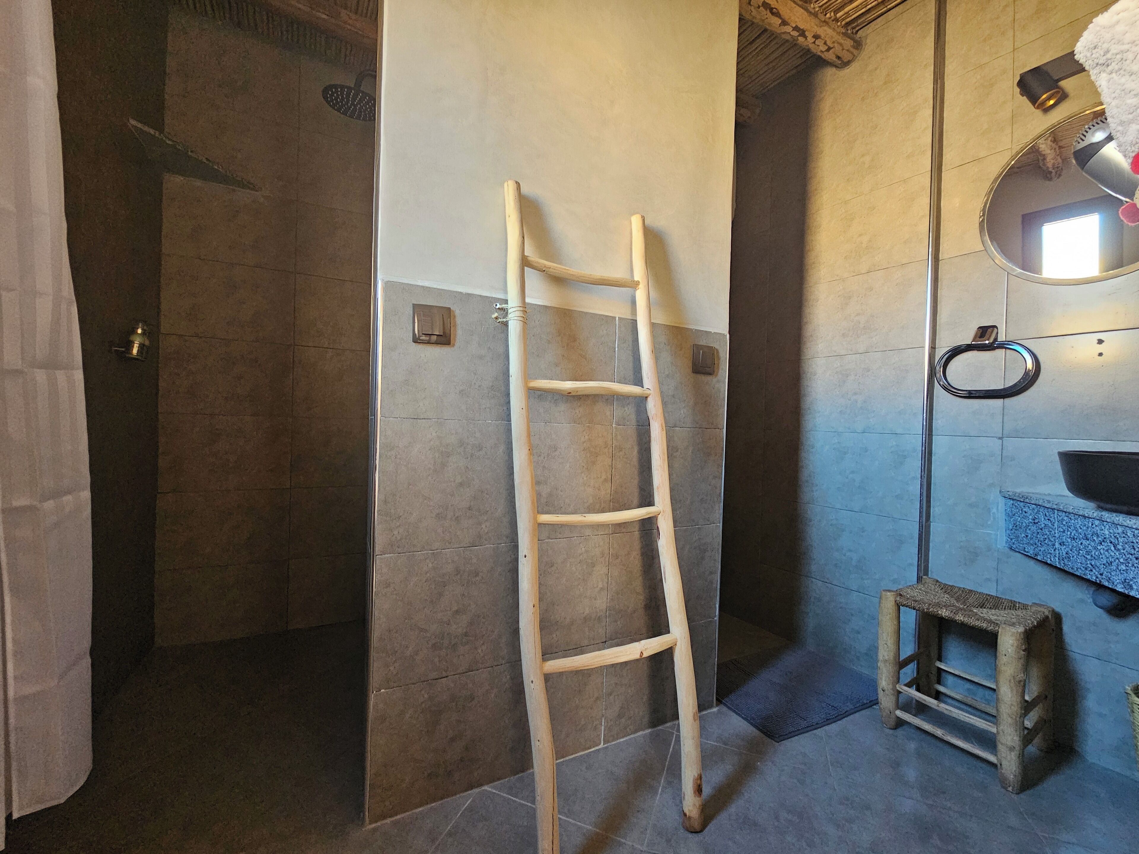 A bathroom with a shower, a wooden ladder, a small stool, a sink, and a mirror.