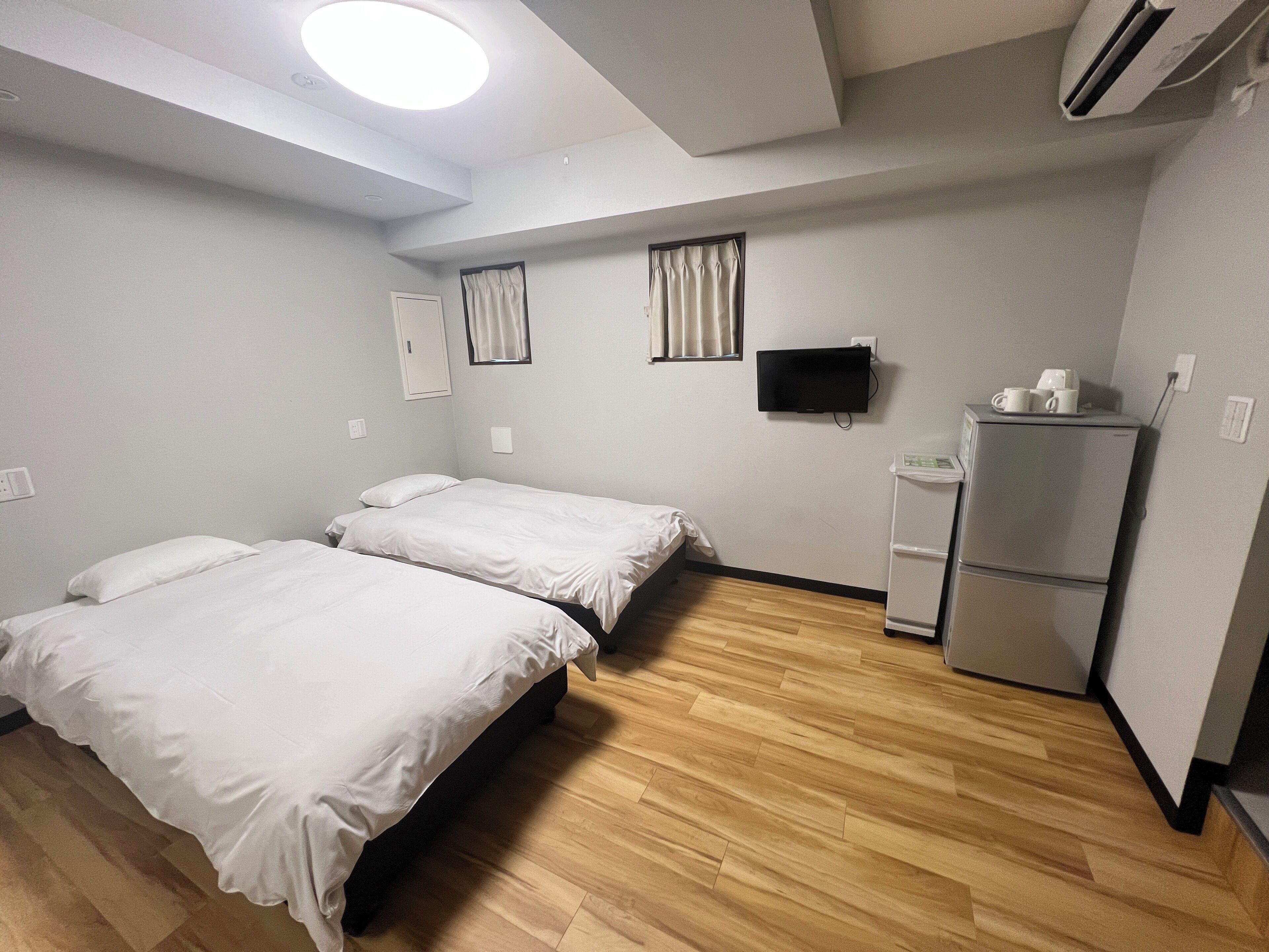 A hotel room with two beds, a flat-screen TV, a refrigerator, and a window with curtains.