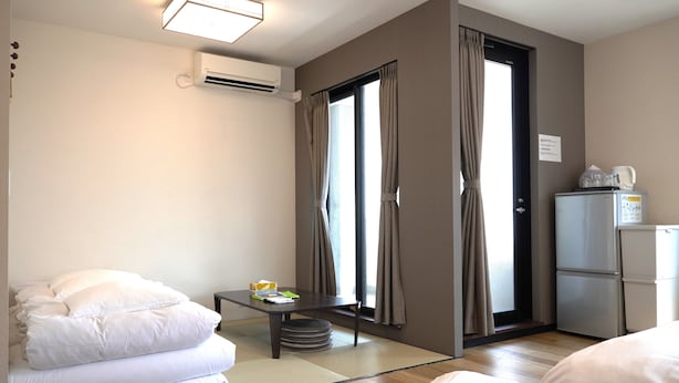 A hotel room with a bed, a small table with a cup and a book, a refrigerator, and a window with curtains. - KYU KYU HOTEL (Tokyo)