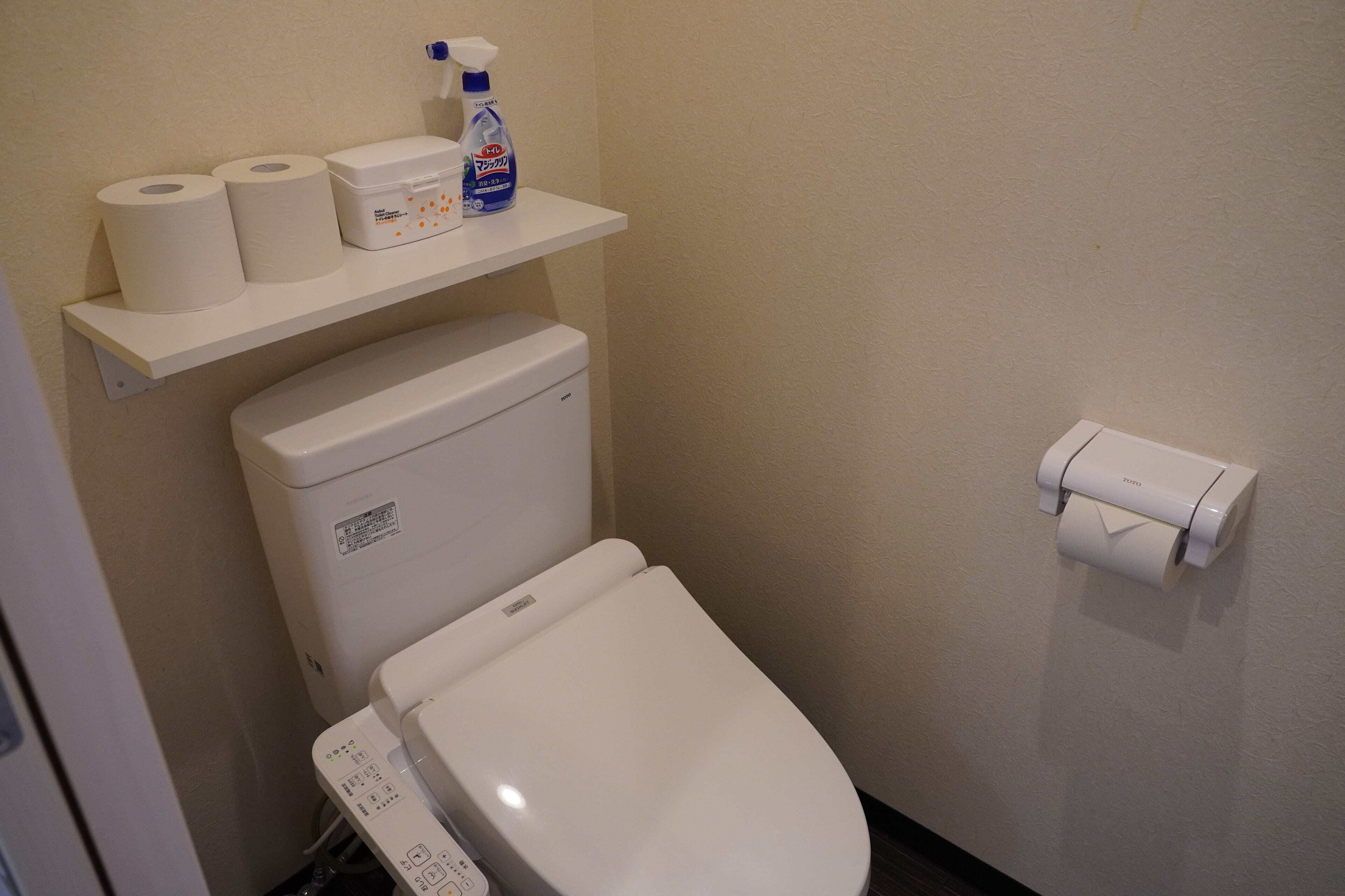A toilet with a control panel, a shelf with toilet paper and cleaning supplies, and a toilet paper holder on the wall.