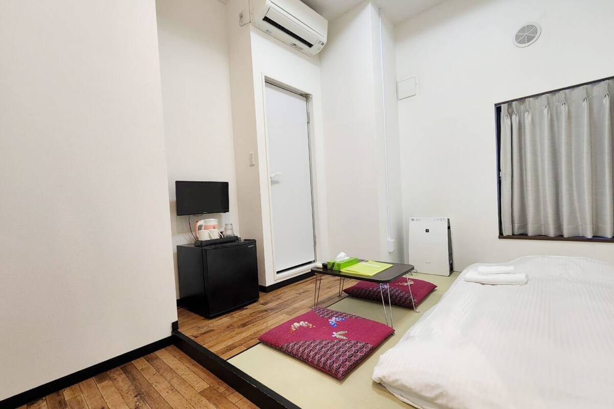 A hotel room with a bed, a desk with a computer, a small refrigerator, a TV, and a window with curtains.