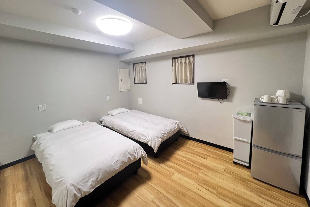 A hotel room with two single beds, a flat-screen TV, a refrigerator, and a window with curtains.