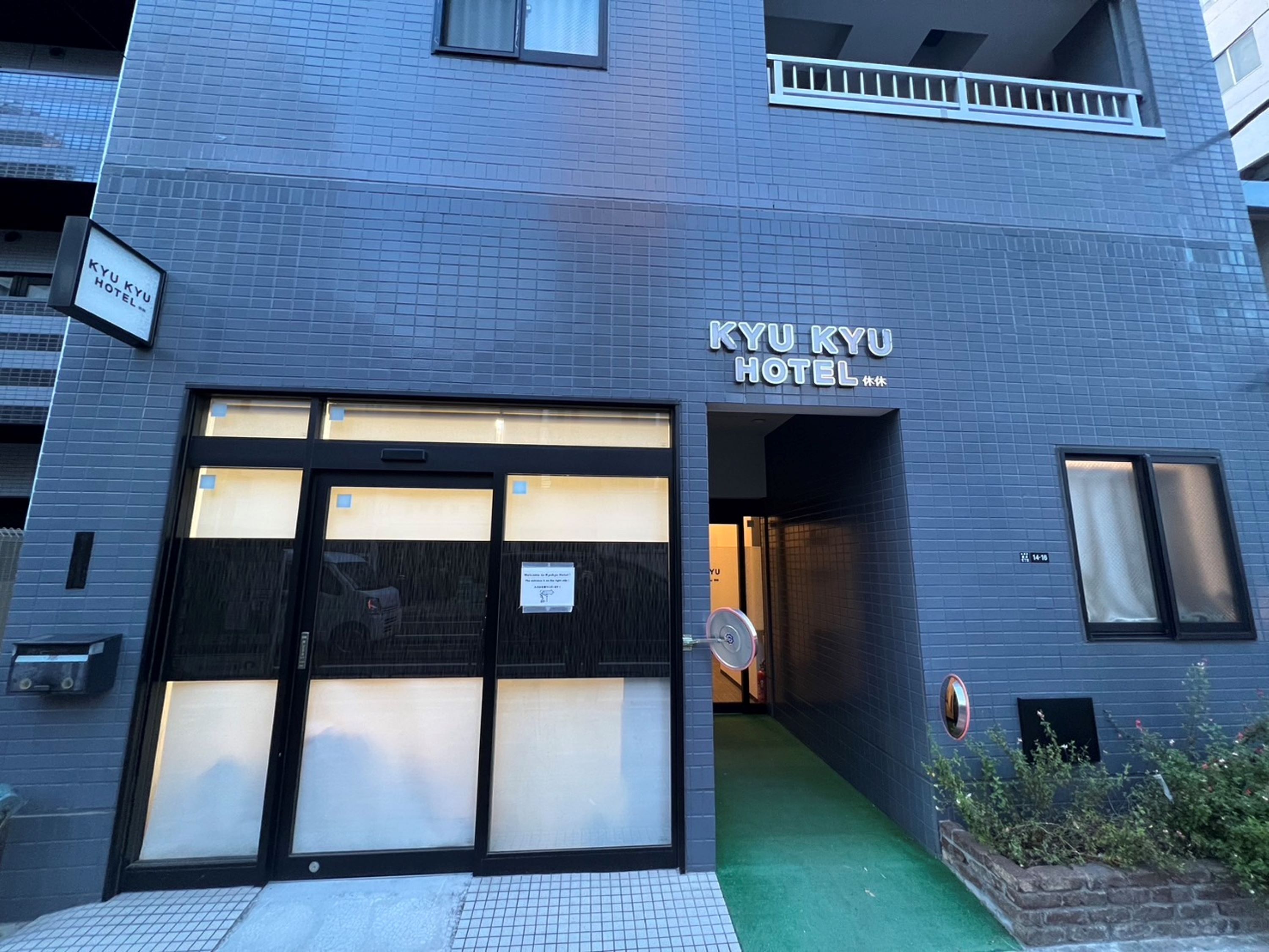 La façade de l’hôtel KYU KYU présente un design moderne, de grandes fenêtres en verre et un espace d’entrée verdoyant.