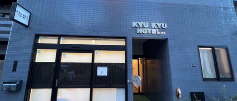 La façade de l’hôtel KYU KYU présente un design moderne, de grandes fenêtres en verre et un espace d’entrée verdoyant.