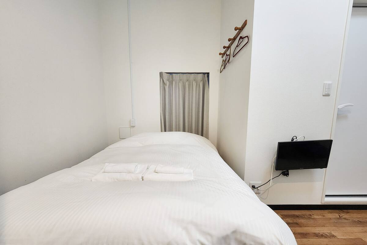 A neatly made bed with pillows, a wall-mounted TV, and a coat rack with coats.