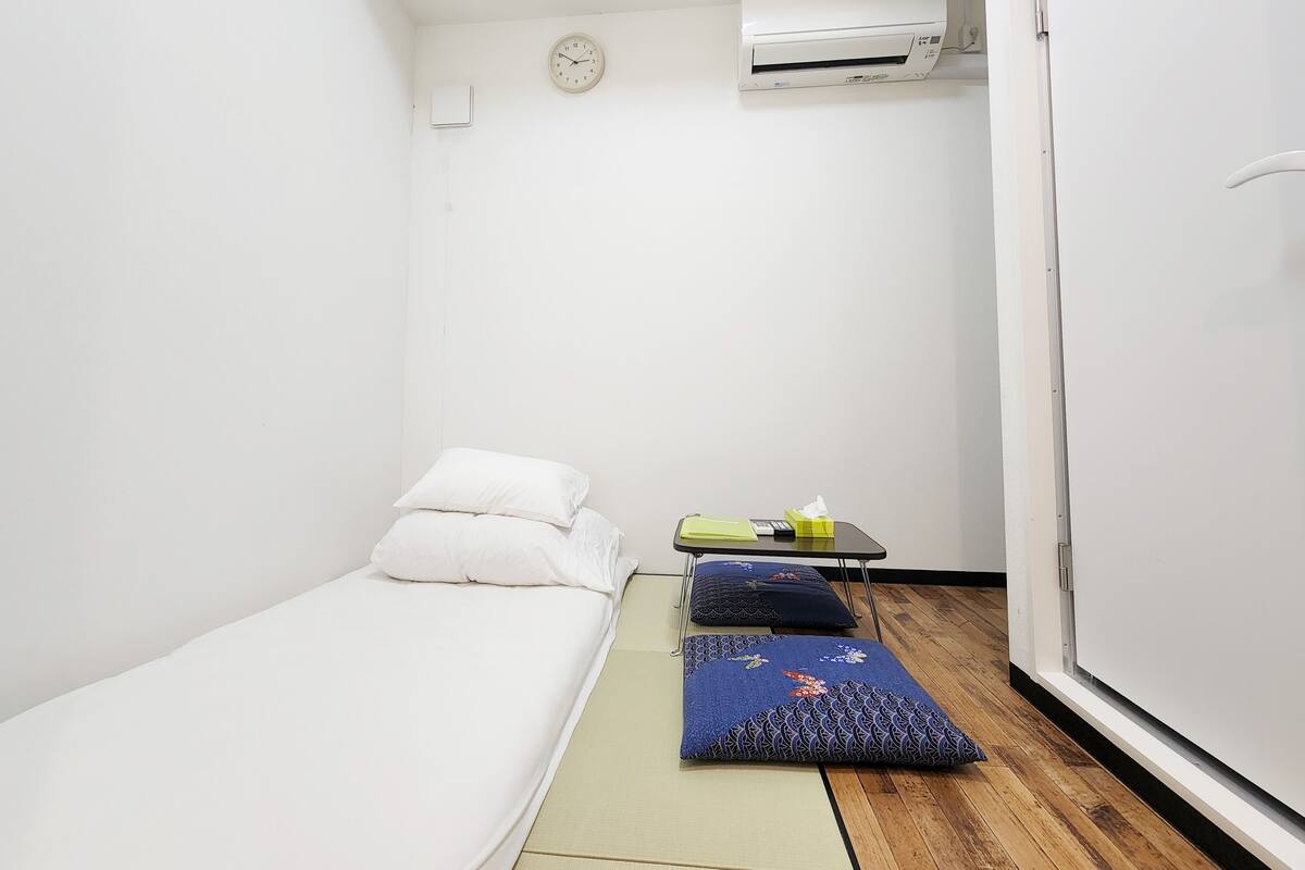 A small, clean room with a single bed, a bedside table with a tissue box and a remote, and an air conditioning unit on the wall.